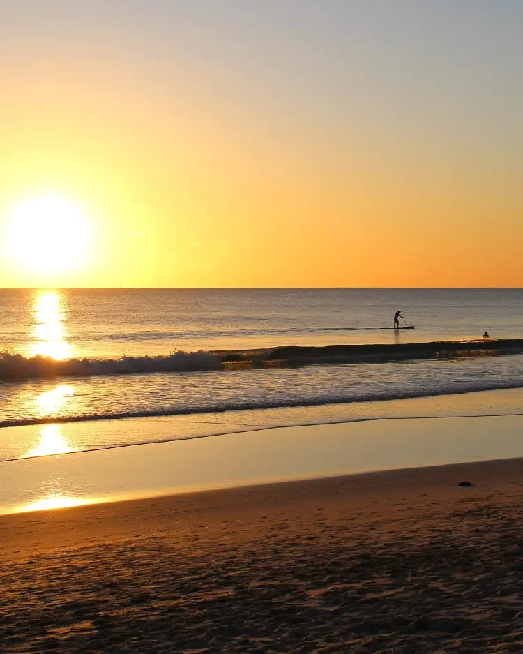 Sunset at Scarborough Beach in Perth and a person doing SUP. Learn about beaches in Perth compared to Brisbane.